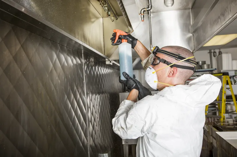 Technician in coveralls spraying degreaser onto the inside face of a stainless exhaust canopy