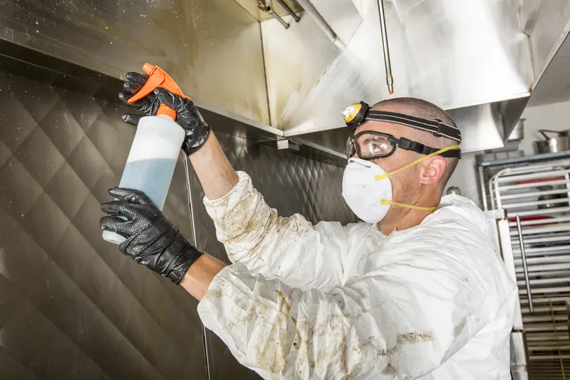 Technician spraying cleaner onto a quilted stainless kitchen wall beside a sheet pan rack